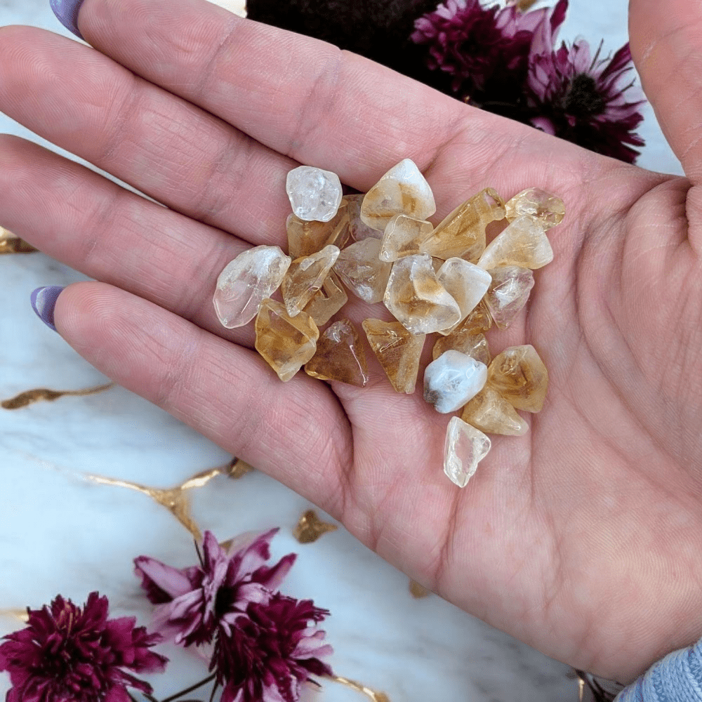 Hand holding small citrine crystal stones with a decorative background of flowers and gold leaf.