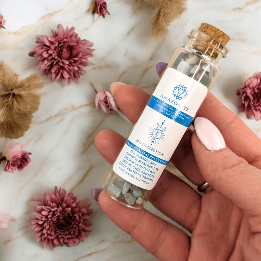 Hand holding a small glass bottle with a cork lid on a marble surface with flowers and amazonite crystals for throat chakra