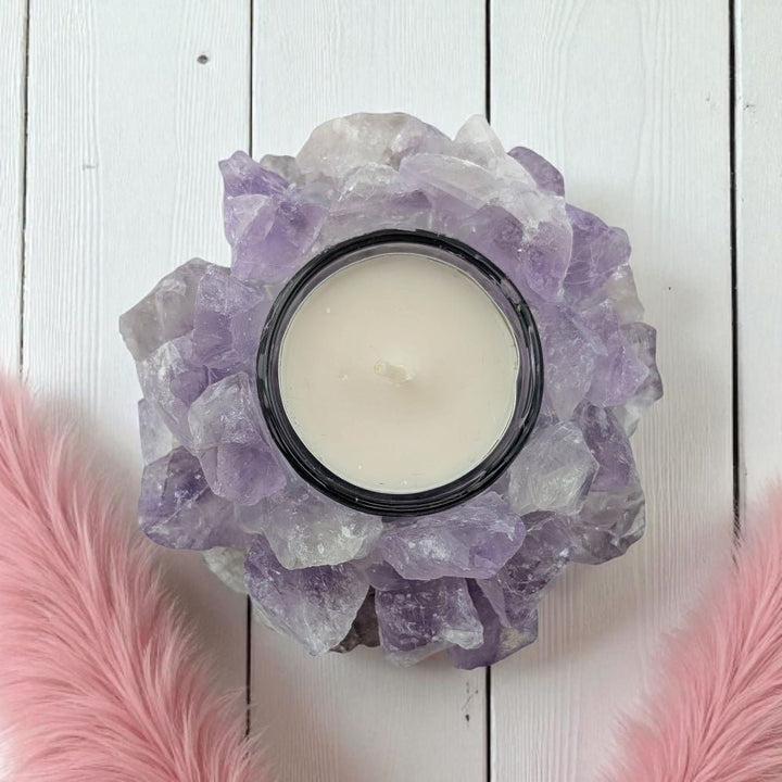 Candle in a decorative holder made of amethyst crystals on a white wooden surface with pink feathers.