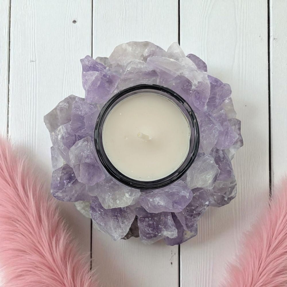Candle in a decorative holder made of amethyst crystals on a white wooden surface with pink feathers.