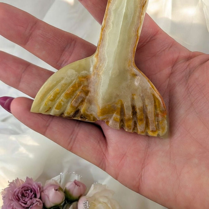 Hand holding a banded calcite mermaid tail crystal with a unique pattern, surrounded by flowers on a light background