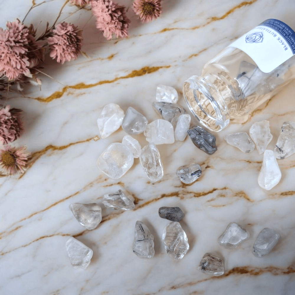 Black Rutilated Quartz stones and a bottle on a marble surface with pink flowers.