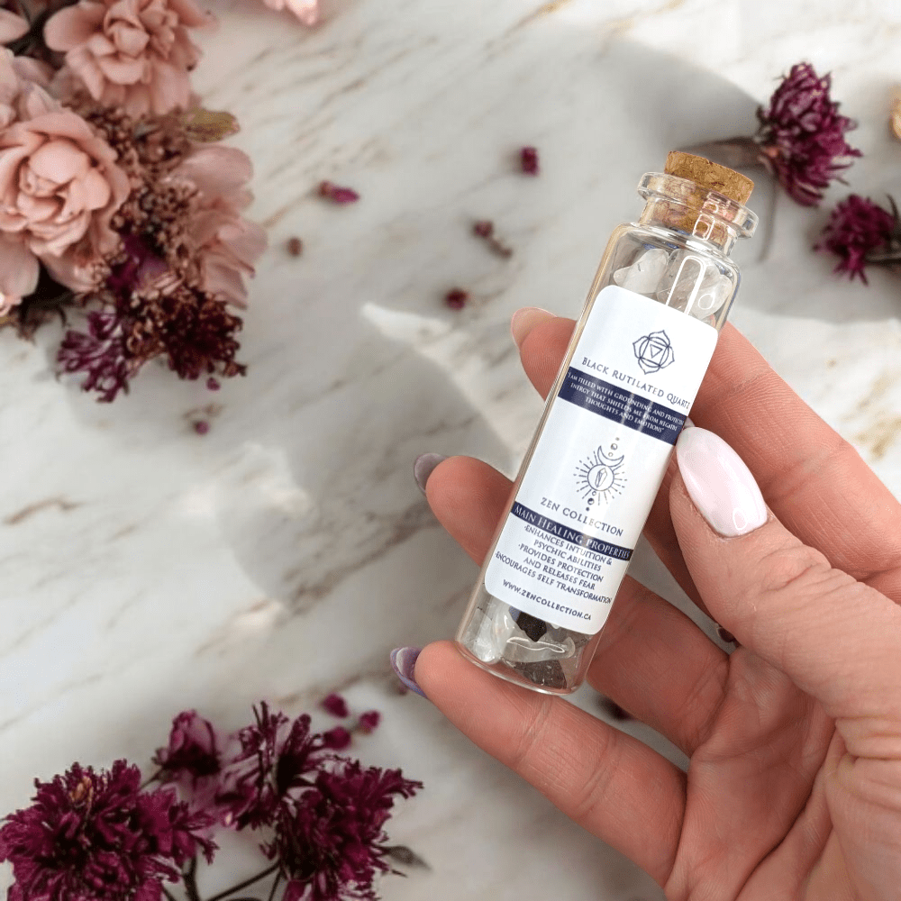 Hand holding a small bottle of  Black Rutilated Quartz crystal chips with a label, surrounded by flowers on a light surface