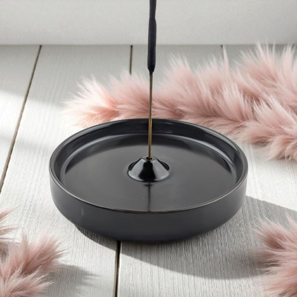 Black incense burner with a single stick of incense on a light wooden surface with pink decorative elements.