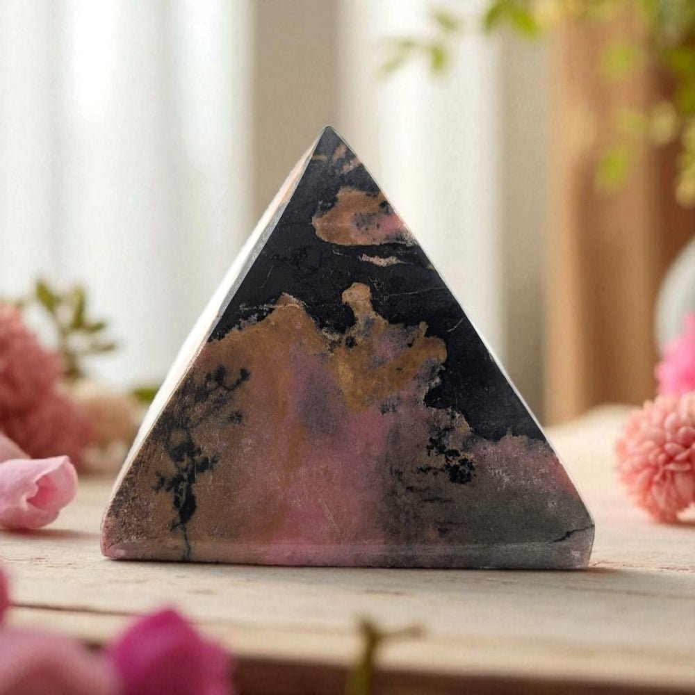 Triangular crystal with pink and black patterns on a wooden surface with blurred pink flowers in the background