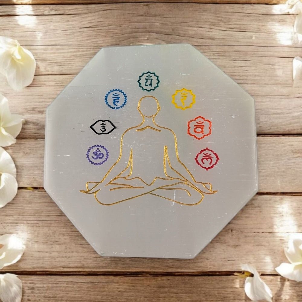 Octagonal selenite crystal with chakra symbols and a human figure on a wooden surface with white flowers.