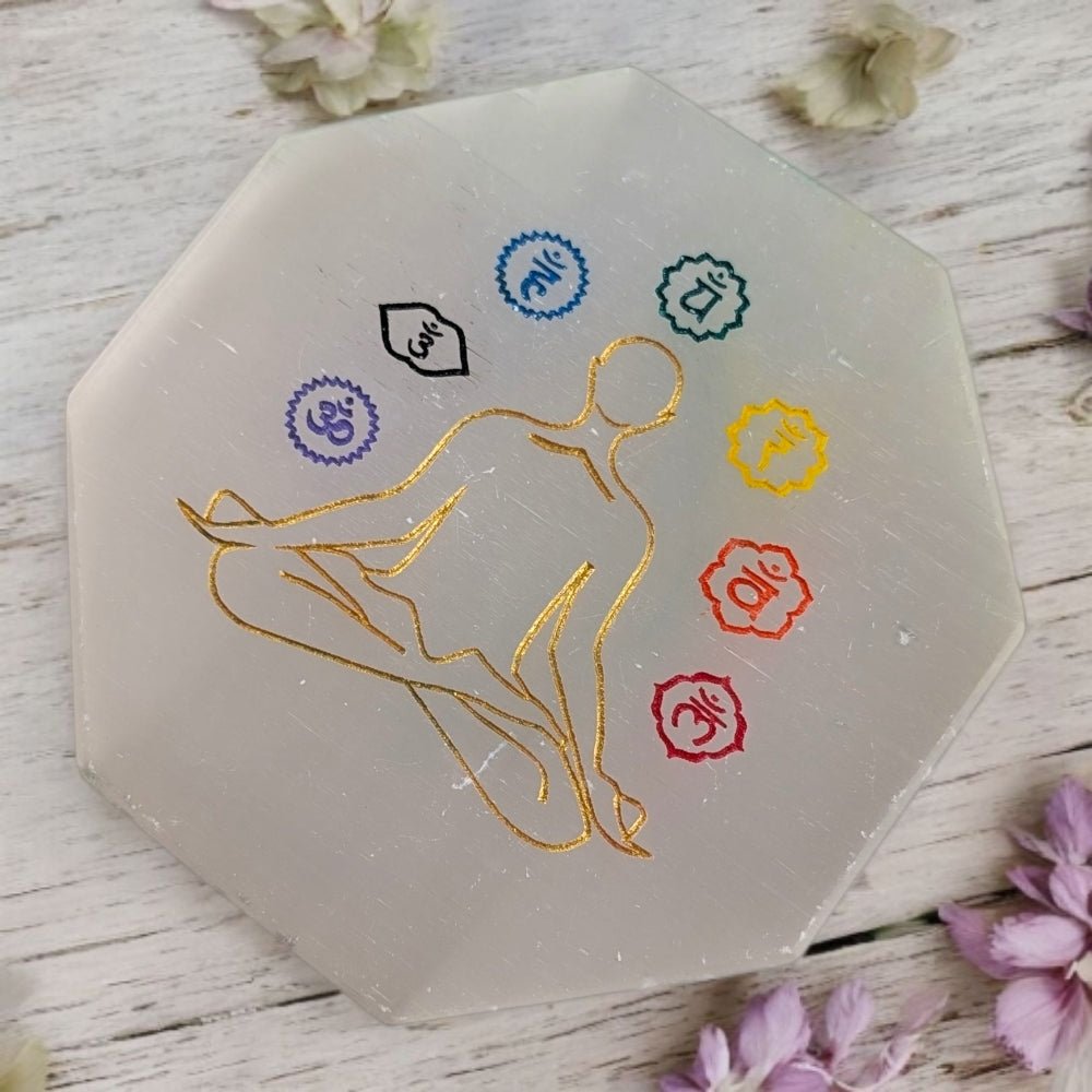 Hexagonal selenite crystal cleansing plate with gold silhouette and chakra symbols on a wooden surface with flowers.