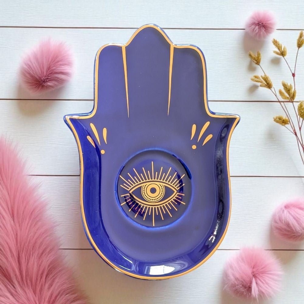 Blue Hamsa hand-shaped plate with gold eye design on a white wooden surface with decorative elements.