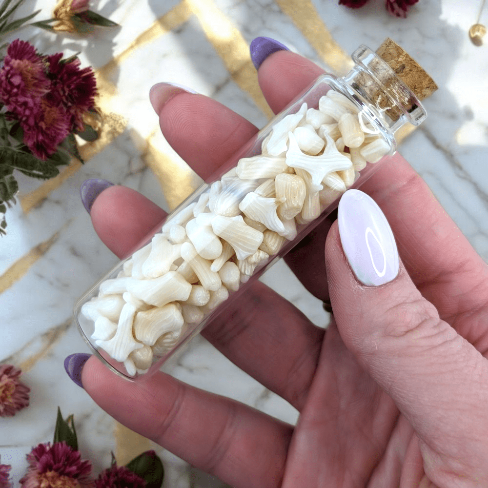Hand holding a small glass bottle filled with agatized coral stones and shells on a marble surface with flowers.