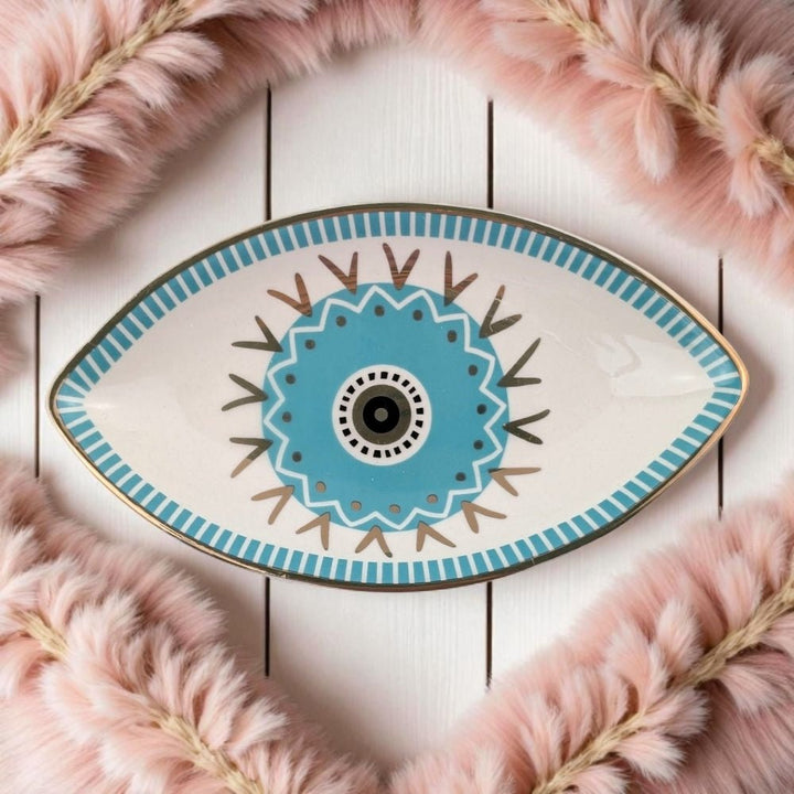 Decorative plate with eye design on a white wooden surface with pink feathers