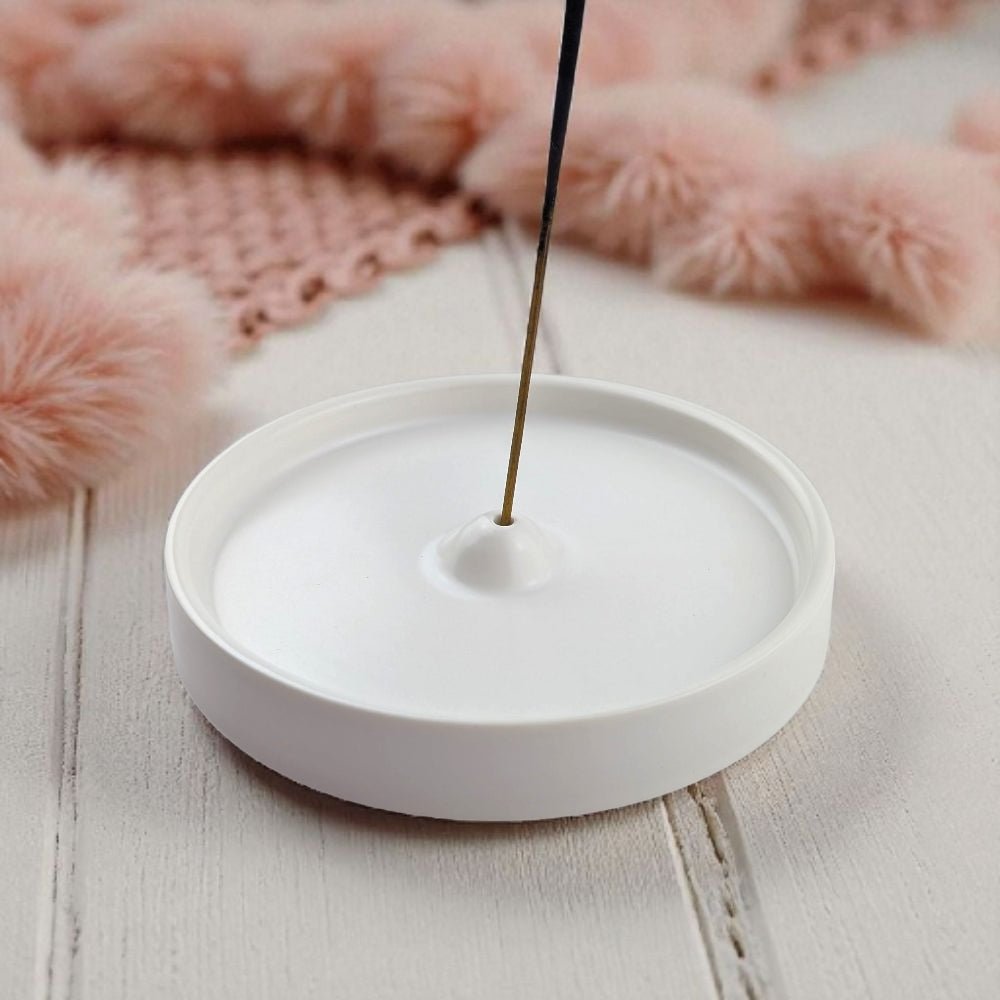 White ceramic incense burner with a single incense stick on a light wooden surface with pink decorative elements in the background.