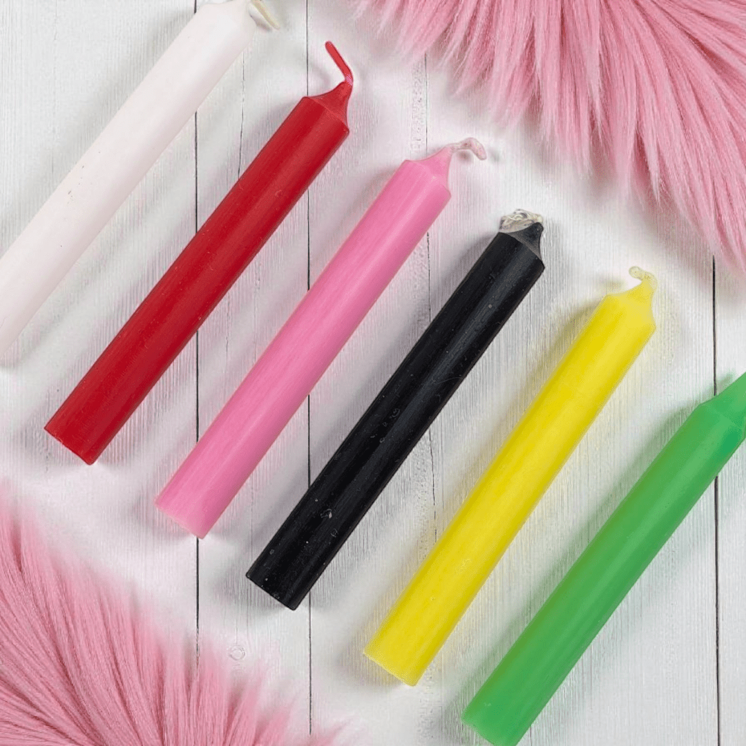 Five colorful candles (red, pink, black, yellow, green) on a white wooden surface with pink decorative elements. Available at Zen Collection Crystal Store in Barrie
