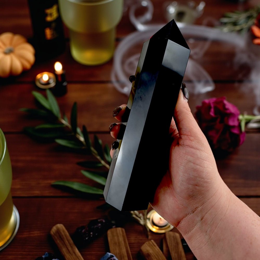 Hand holding a Black Obsidian crystal  Tower with a blurred background of candles and pumpkins on a wooden table available at Zen Collection crystal store near you