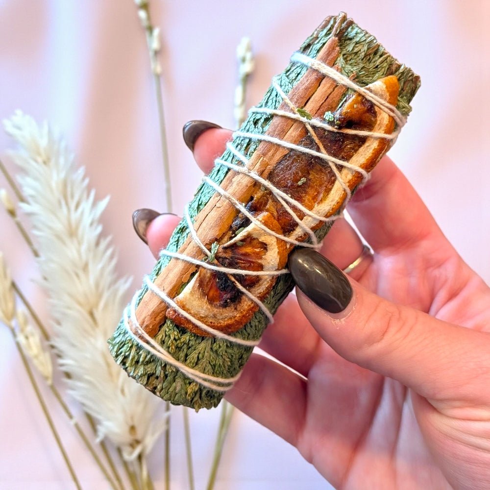Hand holding a bundle of cedar and cinnamon sticks wrapped with twine against a blurred natural background. Available at zen collection crystal store in Barrie