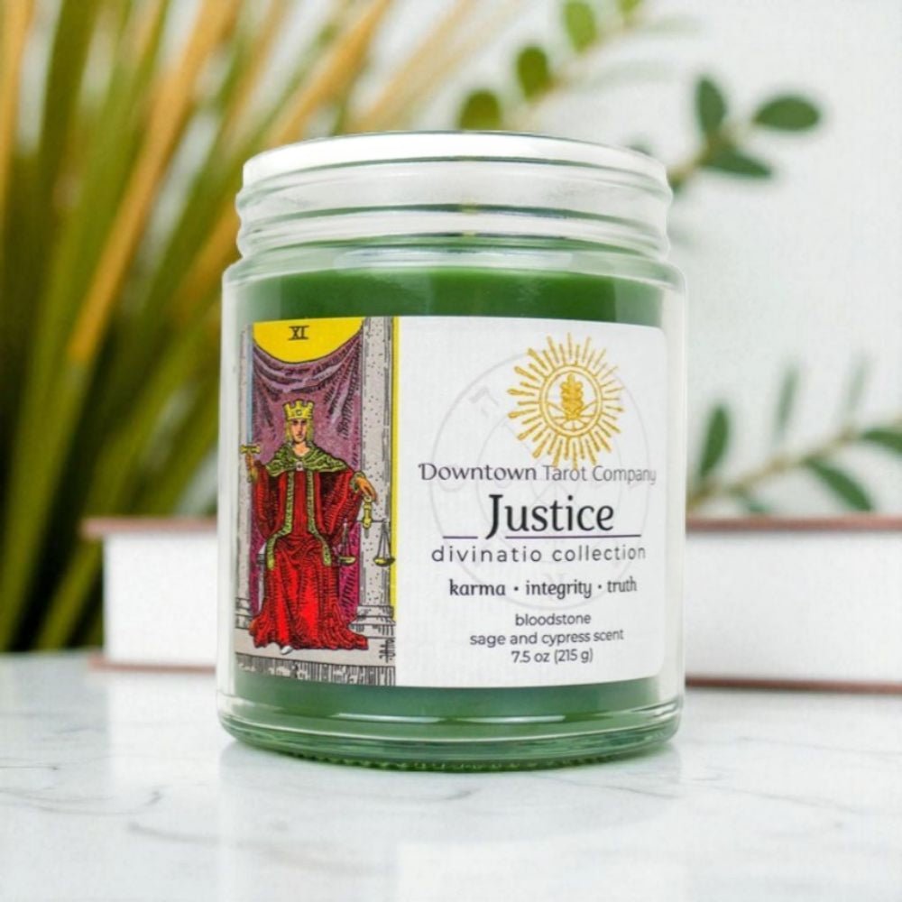 Green justice tarot candle  in a glass jar with a label on a white surface, surrounded by plants available at zen collection metaphysical store
