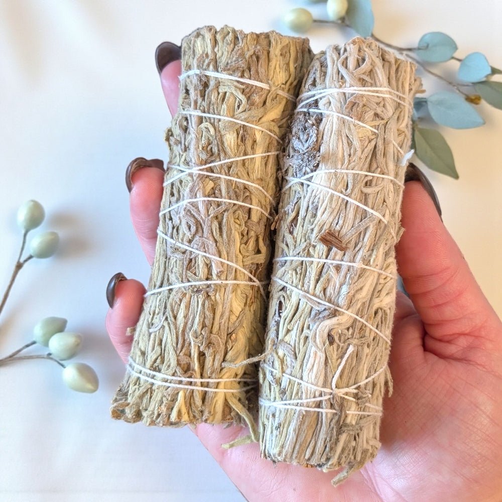 Two bundles of blue sage wrapped in twine held by a hand against a light background with greenery. available at zen collection crystal store