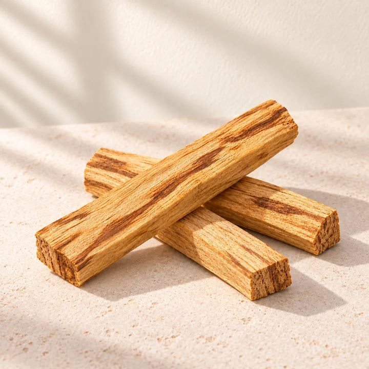 Three unwrapped Palo Santo sticks stacked on cream stone against an ivory background; sold individually.