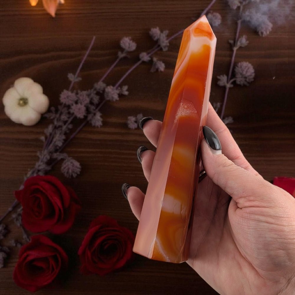 Hand holding a striped orange  carnelian tower for the sacral chakra. surrounded with flowers and roses on a wooden surface. photo is taken by zen collection crystal store near me